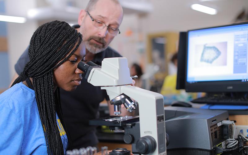 Woman in a lab looking though a mircoscope while the professor adjusts the microscope