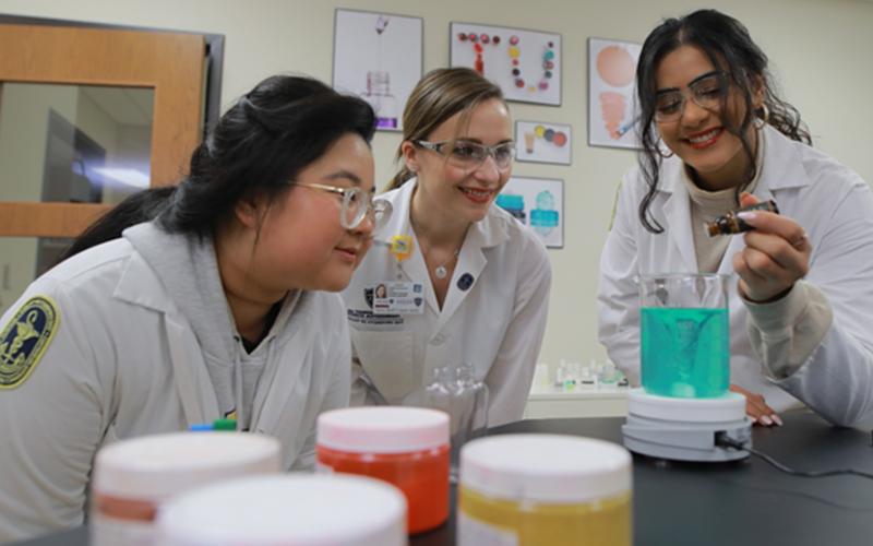 three students in lab coats and goggles watch as one student adds drops into a beaker