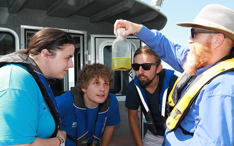 4 people on a boat all wearing life jackets the professor is holding up a bottle of liquid while the three students closly observe the bottle the professor is holding