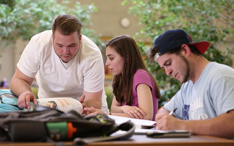 three students at a table , two of them are looking at the same textbook togther the third student is writing things down on paper