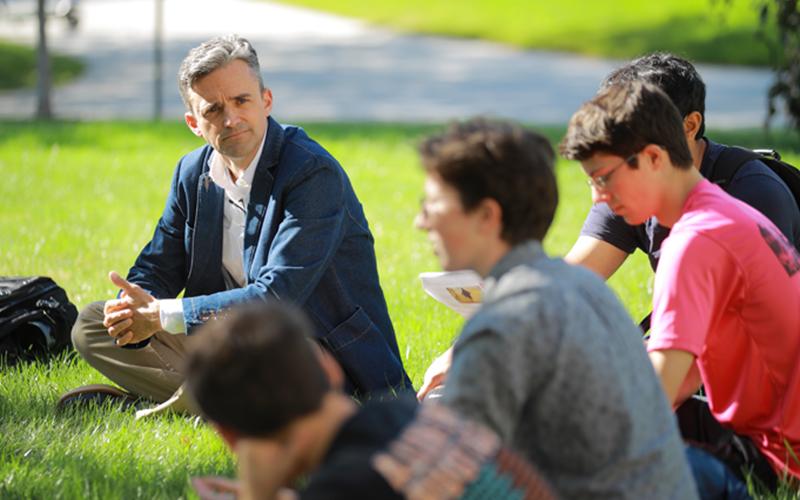 a professor listens in on group discussion during a lecture taking place outside in a grassy patch