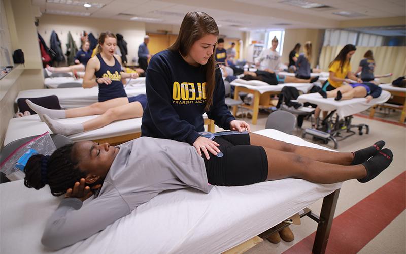 in a room aligned with medical beds, physical therapy students are divided in pairs as one lays on the bed and the other takes measurements