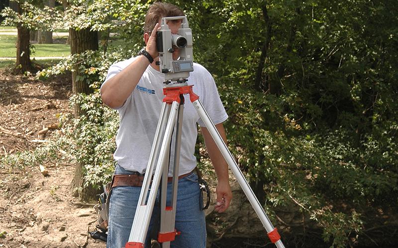 man in a wooded area looks through a large camera device 