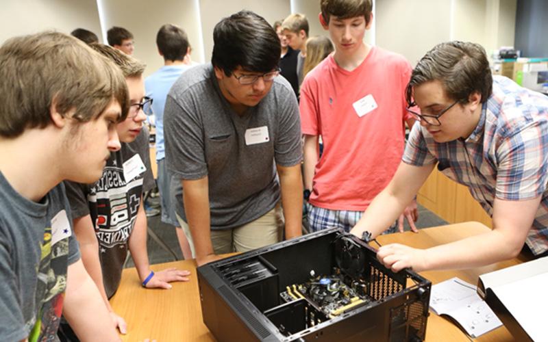 a group of student stand huddled around a table and observe an unassembled computer
