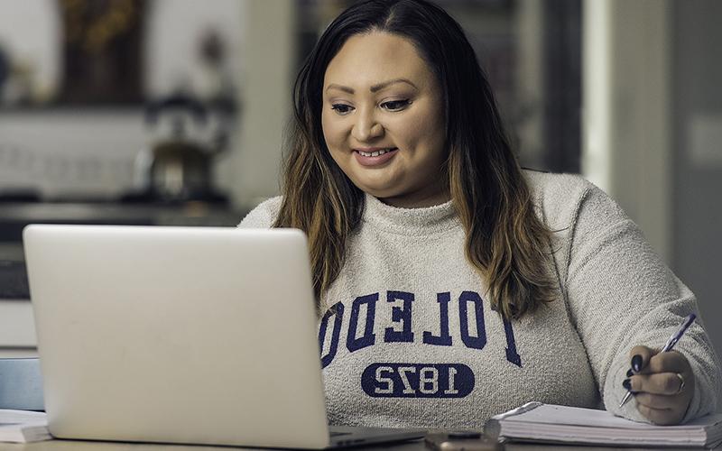 woman wearing a UToledo sweatshirt looks down at her open laptop next to her pen and notebook